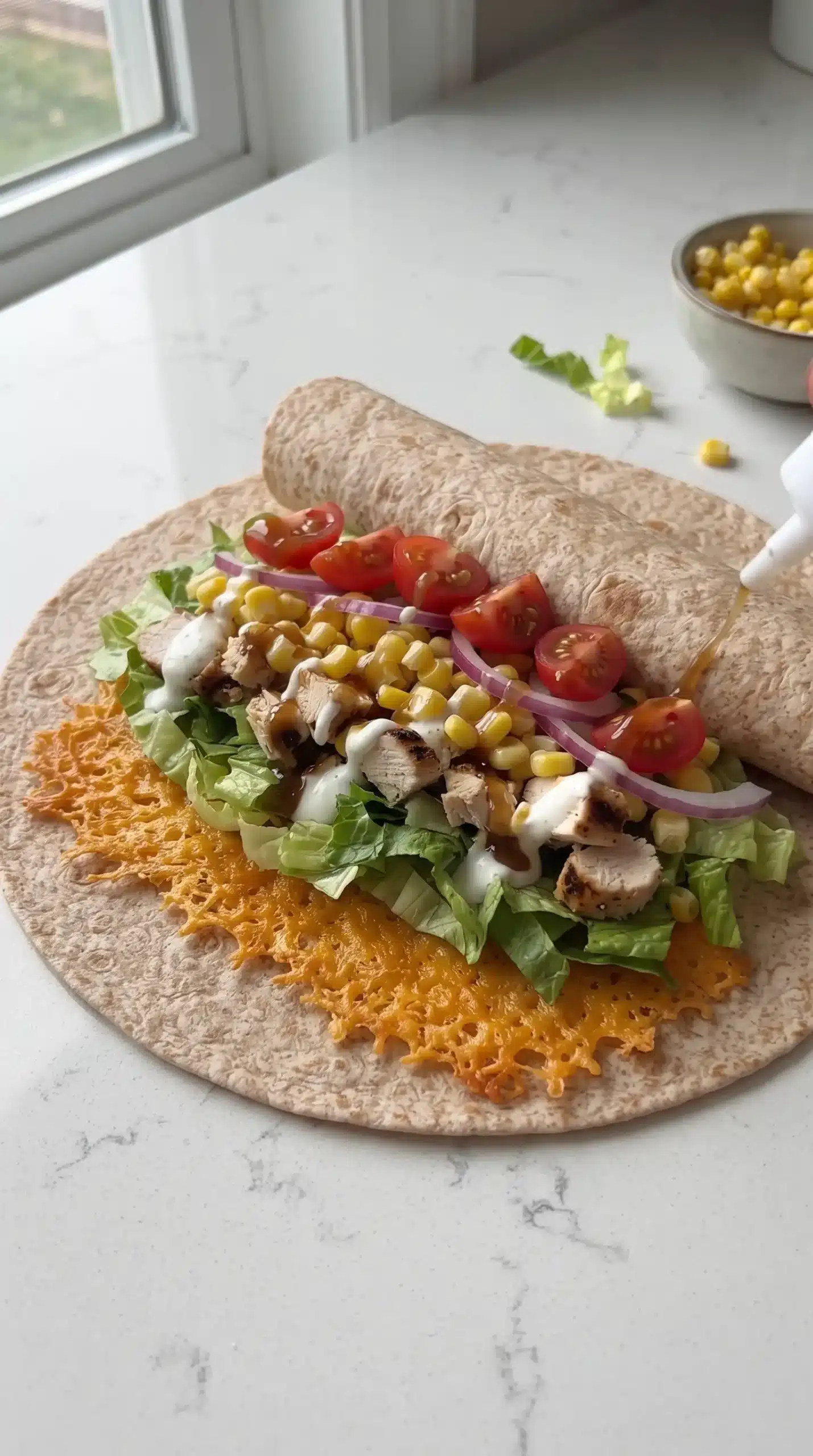 Close-up preparation shot of Ang's Chicken Wraps being assembled on a whole wheat wrap laid flat on a clean neutral countertop. Show the layering process in progress: a cooled cheddar cheese crisp aligned along one edge of the wrap, small pieces of grilled chicken, fresh corn kernels, sliced tomatoes, shredded lettuce, thin red onion, and controlled drizzles of ranch dressing and sweet tangy sauce being added before folding. Keep the wrap partially open to clearly show the organized filling layers, realistic ingredients, soft natural light, minimal props, and no fully finished plated wrap.