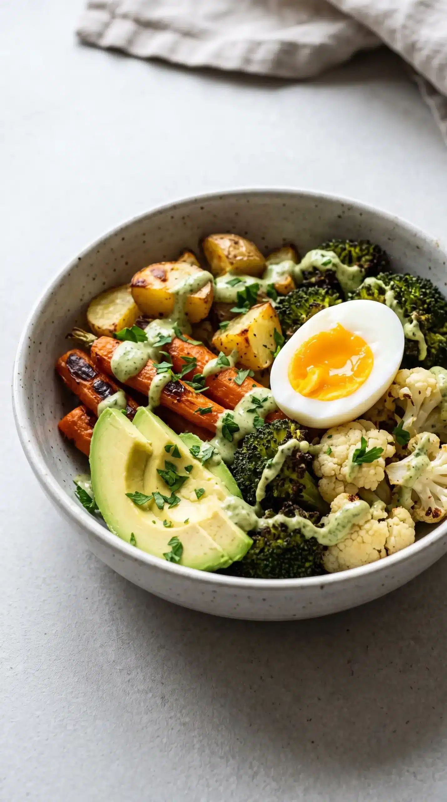 Roasted vegetable bowl topped with green tahini, avocado, fresh herbs, and a halved hard-boiled egg