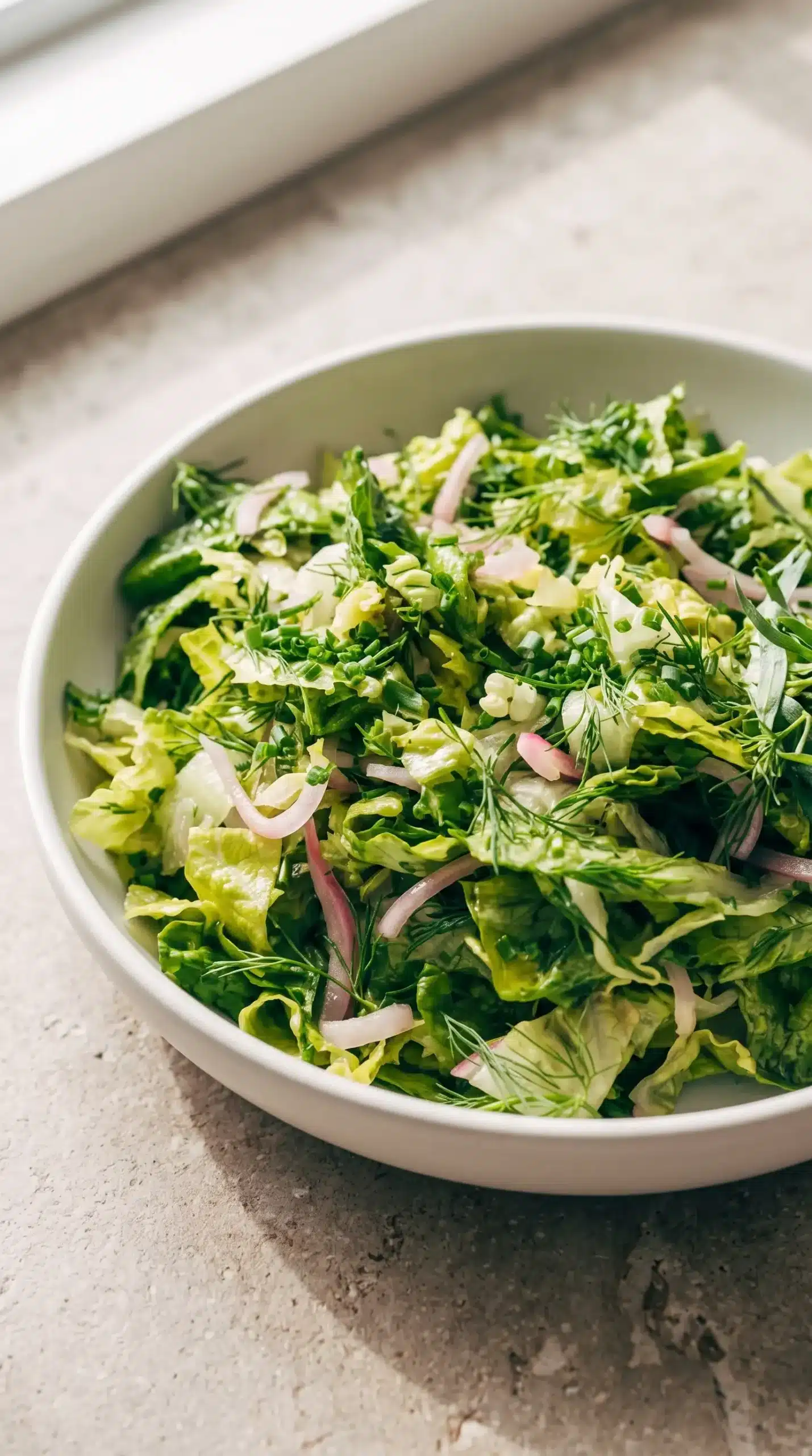 Professional food photography close-up of a finished Liz's Bistro Salad in a single shallow white ceramic bowl, featuring crisp chopped romaine, torn butter lettuce leaves, fresh green tarragon, dill, and chives with thin pink pickled red onion slices distributed throughout. The salad should look naturally tossed with a light lemony dressing coating the leaves, fresh and balanced rather than overloaded. Place the bowl on a simple neutral stone-colored surface with soft natural daylight, realistic textures, a close three-quarter angle, minimal styling, no extra servings, no wooden table, no decorative props.